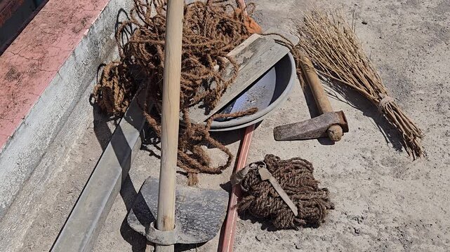 Construction tools for traditional plastering: a mixing bowl, shovel, leveler straight edge, & hammer chisel axe are scattered. Coco coir rope is piled high, used for lashing bamboo framing for stands