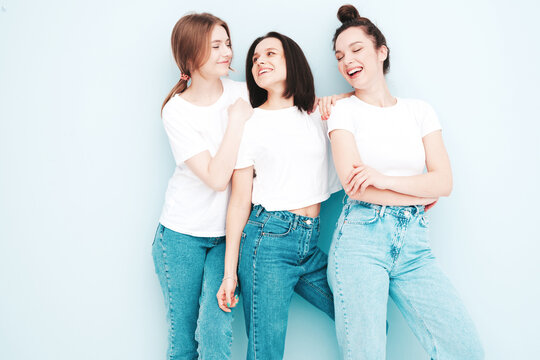Three young beautiful smiling hipster female in trendy same summer white t-shirt and jeans clothes. Sexy carefree women posing near light blue wall in studio. Cheerful and positive models having fun