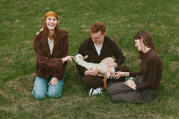Naklejka premium A warm outdoor scene features three friends kneeling on grass, gently petting a curious goat kid, capturing a lifestyle moment of friendship, care, and playful bond with animals in nature.