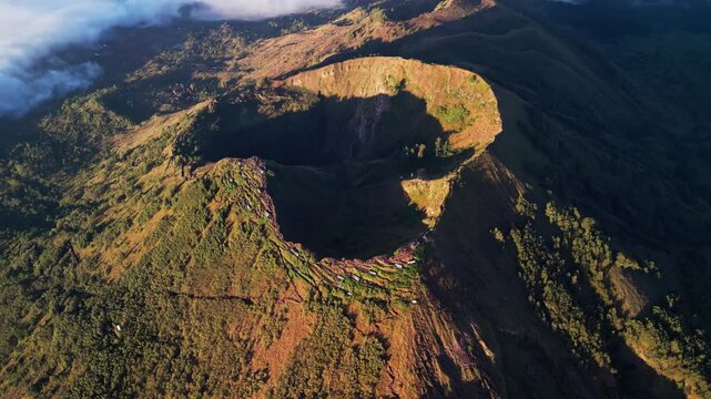Volcanic terrain with deep caldera and steep slopes under golden light highlighting rugged mountain and surrounding forested landscape in wide aerial drone view