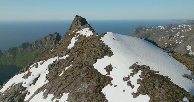 Aerial drone flight over Munken summit in Lofoten, Norway, as two hikers cross a snow patch atop a rugged granite mountain under clear skies.