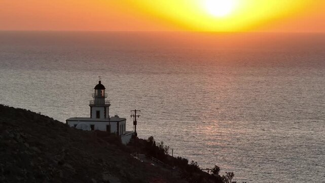 Drone revealing the Santorini lighthouse during golden hour at sunset, capturing the horizon, caldera cliffs, and the deep blue Aegean Sea in warm light.