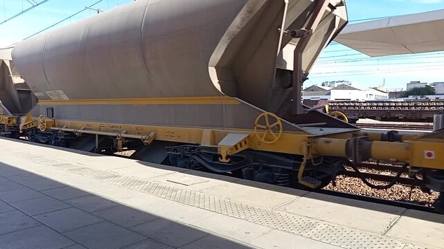 Freight train hopper wagon at a railway station platform, showing industrial rail transport for bulk materials with steel structure, unloading design, and electrified infrastructure in the background.