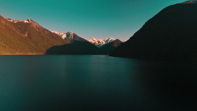 Deep dark pristine lake waters gently rippling extending toward spectacular rugged alpine mountains featuring bright white snow patches glowing from golden evening sunlight high altitude approach air