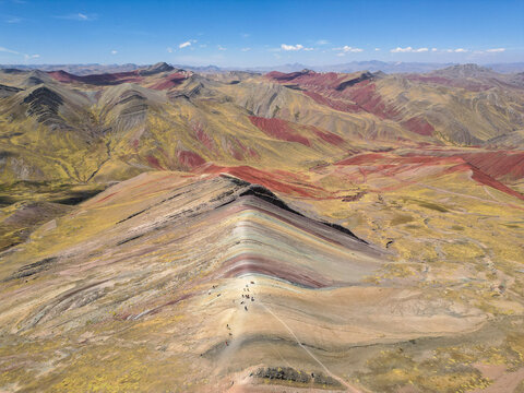 Aerial view of Vinicunca Rainbow Mountain showing vibrant mineral stripes and hikers on the ridge among rugged Andean peaks in Quispicanchi Province, Cusco, Peru.