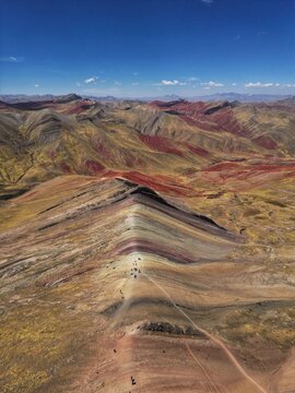 Aerial view of Vinicunca Rainbow Mountain showing vibrant mineral stripes and hikers on the ridge under a clear blue sky in Quispicanchi Province, Cusco, Peru.