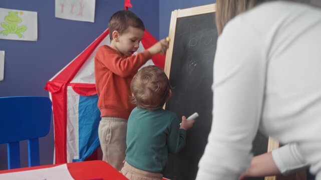 Young boy drawing on blackboard with chalk, pointing finger while toddler scribbles beside him in building; curiosity cooperation.