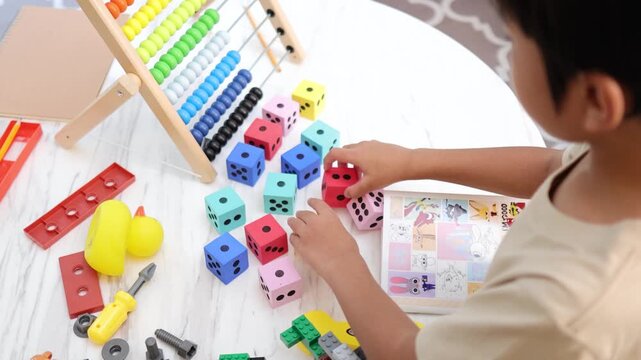 Asian little boy playing with colorful wooden dice and educational toys on table at home.