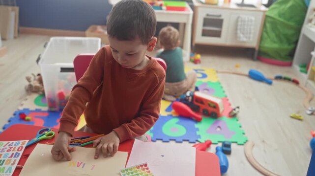 Boy arranging colored pencils on a small table, fingers on paper while drawing, toys and foam number mat scattered on floor in a building; concentration sibling play.