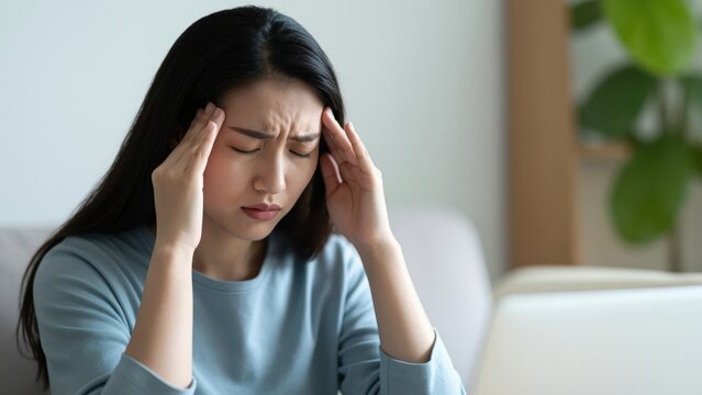 Woman with Headache: An asian woman massages her temples in distress, experiencing a tension headache after prolonged screen usage.