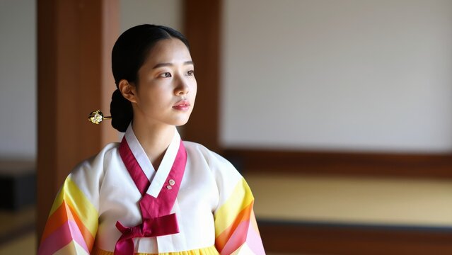 Elegant Korean Woman in Traditional Attire: A serene young woman, adorned in a vibrant, multi-colored hanbok, gazes thoughtfully into the distance within a traditional Korean interior.