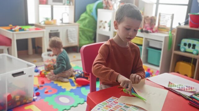 Boys at table with colored pencils and paper, hand folding paper while one brother plays with blocks in a building; quiet concentration.