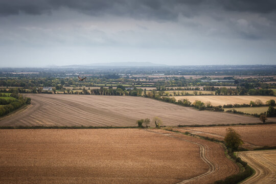 Aerial view of Keogh's Farm with harvested agricultural fields, a bird of prey in flight, and distant hills under a moody overcast sky in Oldtown, County Dublin, Ireland.