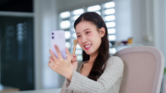 Asian women beauty blogger influencer recording makeup tutorial video using smartphone holding brush sitting home office natural light modern interior cheerful