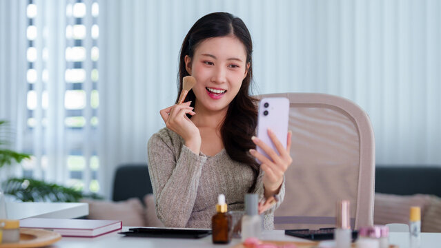 Asian women beauty blogger influencer live streaming makeup tutorial video call using smartphone camera recording beauty content sitting desk home office
