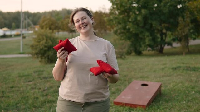 Portrait of happy woman showing red corn bags for cornhole game to camera. Player enjoying quality of sports equipment, smiling to camera before successful outdoor game in park.