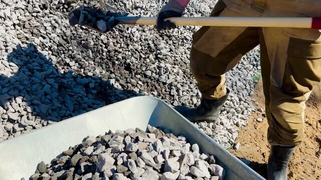 An unrecognizable man loads crushed stone into a construction wheelbarrow using a shovel
