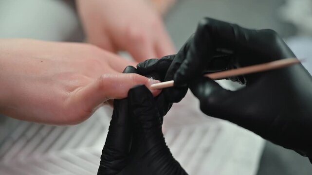 A person uses a wooden stick to push back cuticles on a client's hand. The client appears to be an adult female with fair skin, sitting in a salon setting while receiving a manicure
