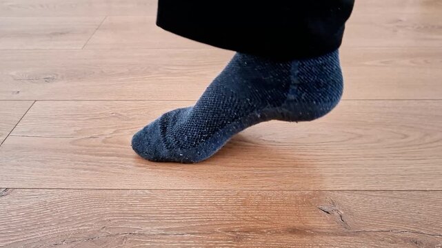Foot in dark blue textured sock balances on wooden floor with heel lifted, showing tiptoe stance, motion, and posture related to exercise, dance, or everyday movement