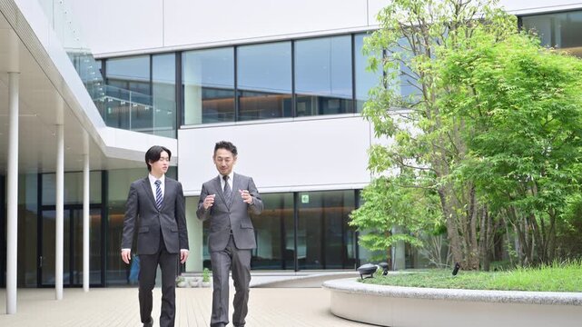 A video showing two salesmen in suits walking and talking in front of a building outdoors; a scene of a subordinate and a supervisor having a conversation