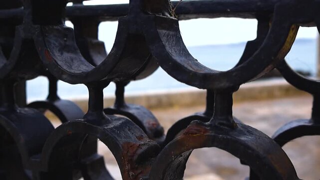 Close up on a rusty black wrought iron fence with a shallow depth of field, revealing the blurred sea, the beach, and the coastline of the Atlantic Ocean in the background