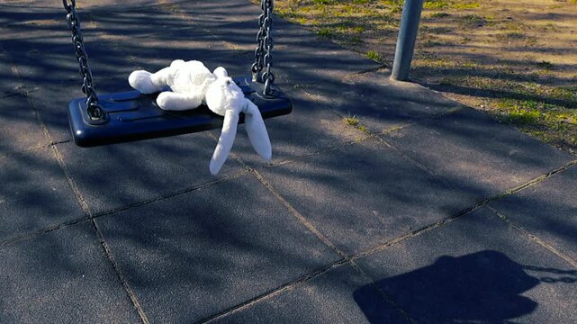 Sad forgotten white stuffed bunny toy swinging all alone on an empty swing set at a desolate playground, symbolizing loneliness, lost childhood, neglect, and abandonment