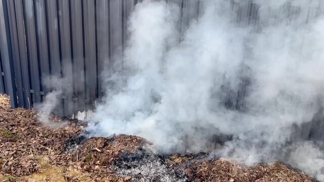 Dry leaves burn and smolder near a fence on a countryside property, producing smoke in an outdoor setting