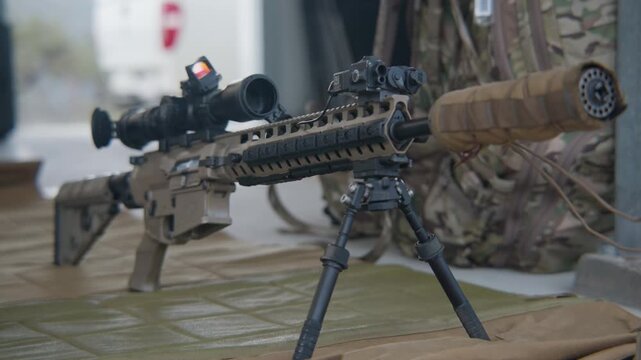 Pan left, A side on view of a sniper rifle set up on a mount that is on display with a camouflage backpack in the background.