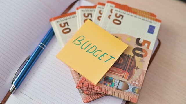 A stack of euro banknotes is shown with a yellow sticky note on top that reads Budget. A pen is placed alongside the money, suggesting organization of finances for planning and tracking