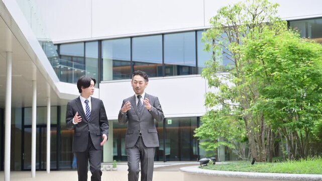 屋外のビルの前でスーツの営業マン2人が会話しながら歩くコミュニケーション　部下と上司が会話する話すシーンから動画　スローモーション