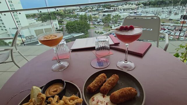 A rooftop bar setting with cocktails and entrees on the table with menus and the city scape in the background.