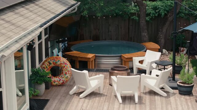 Elevated drone shot of a backyard deck with a hot tub, white Adirondack chairs, donut float, and outdoor TV under string lights. Perfect for outdoor living ads, lifestyle, and home content.