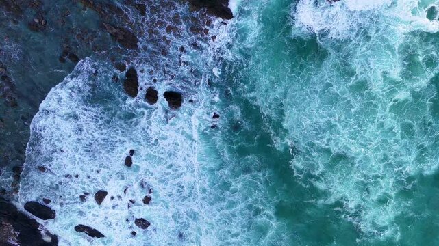 Aerial slow motion view of ocean foam colliding with turquoise water near rocky coast.