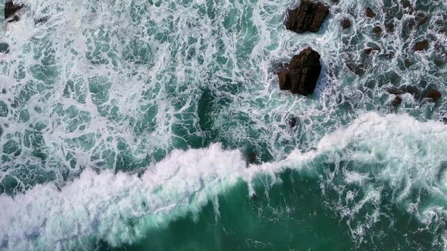 Drone view of a clean wave line rolling toward rocky coastline in turquoise water.