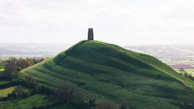 Aerial drone view of Glastonbury Tor with St Michael&rsquo;s Tower standing on top of a lush green hill in Somerset England surrounded by rolling countryside and soft natural light