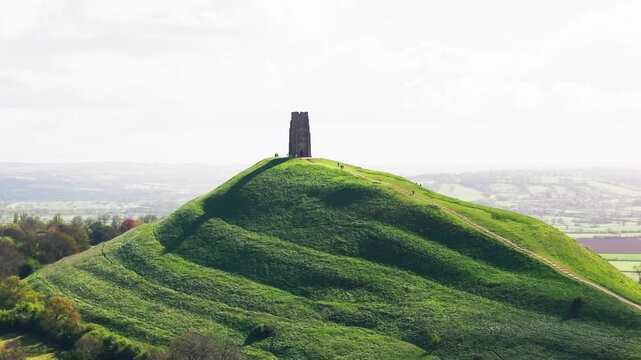 Tight drone shot of Glastonbury Tor highlighting the textured green slopes and footpath leading to St Michael&rsquo;s Tower with distant Somerset landscape