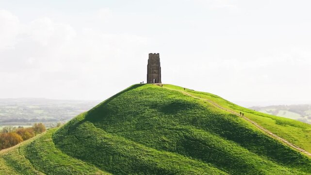 Aerial drone shot of Glastonbury Tor featuring a winding path with hikers ascending a vibrant green hill towards St Michael&rsquo;s Tower in Somerset England