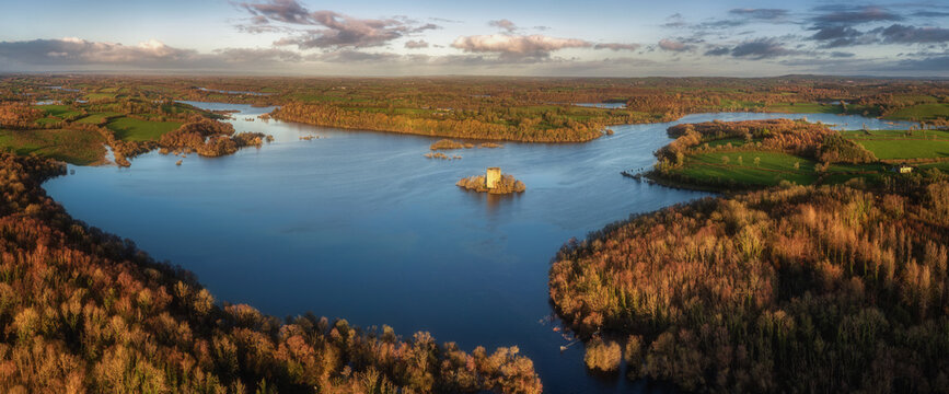 Aerial view of Clough Oughter Castle on a small island in Lough Oughter, surrounded by vibrant autumn forests and calm blue water under a cloudy sky at sunset in County Cavan, Ireland.