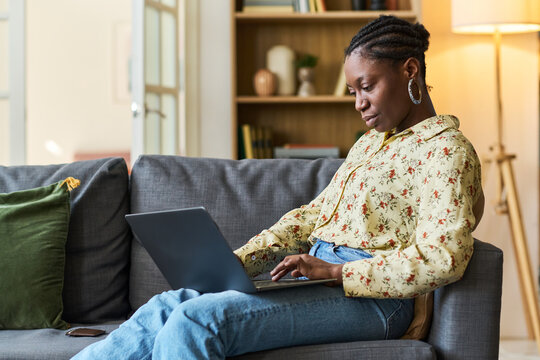 Young adult Black woman using laptop on sofa, planning trip and managing online travel booking from home. Ideal for remote lifestyle, vacation preparation, digital services marketing