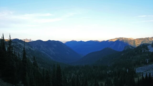 A wide shot of a mountain valley at sunrise, showcasing the rugged peaks of the Tatoosh Range and the lush evergreen forests of the Pacific Northwest. The early morning light casts a golden glow on th
