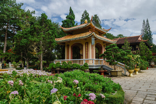 A serene Buddhist temple at Thien Vien Truc lam in Da Lat, Vietnam, with vibrant flowers and manicured bonsai trees. Visitors stroll through the peaceful grounds.