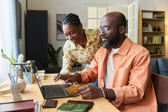 Black couple preparing for trip, booking travel on laptop and holding credit card with passports on desk. Useful for vacation planning, online booking, travel finance marketing