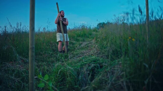 A Man is Standing on a Grassy Hillside, Holding a Long Wooden Pole, Engaged in Farm Chores Under a Clear Blue Sky - Static Shot