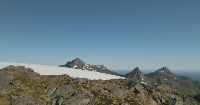 Aerial drone shot of a couple walking across a snow patch on Munken summit in Lofoten, Norway, with granite mountains and a distant frozen lake under a clear blue sky.