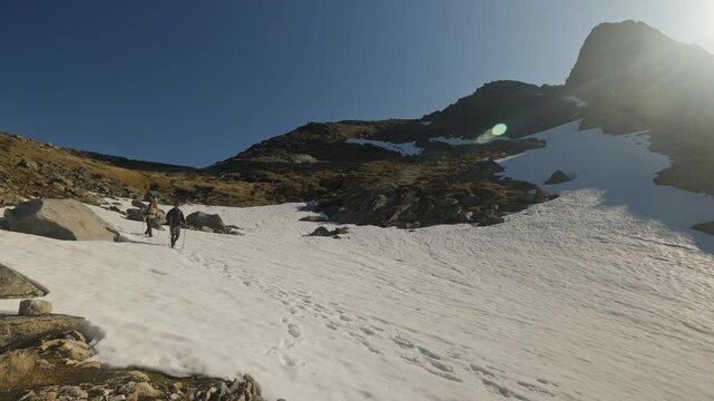 Hikers cross a snowfield in an alpine landscape under a clear blue sky with sun flare, surrounded by rugged granite peaks in Lofoten, Norway.
