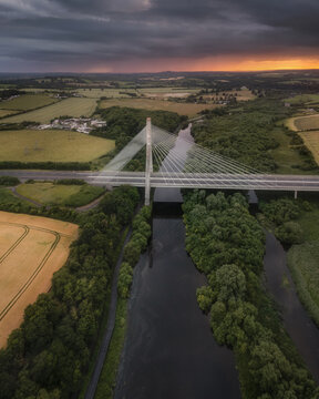 Aerial view of the Mary McAleese Boyne Valley Bridge spanning the river amidst lush green forests and fields under a dramatic sunset sky in Drogheda, County Louth, Ireland.