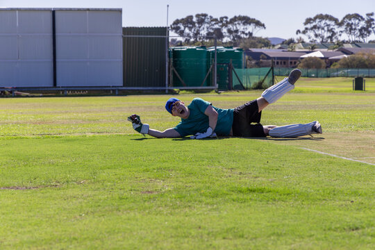 Cricketer lying on grassy cricket field near boundary line wearing pads and gloves by sight screen