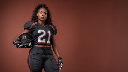Obraz premium Confident African American female tackle football player in a black uniform holds a helmet against a warm brown studio background with ample copy space.