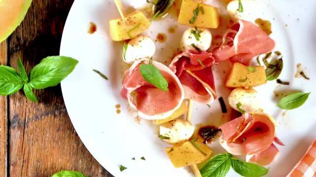 top view of fresh prosciutto and melon skewers with mozzarella balls and basil on a white plate and rustic wooden table