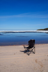 Black camping chair on a sandy beach facing a melting frozen lake. A single folding chair sits on a sandy shore, offering a view of a vast, calm lake with drifting ice fragments © Alex Barera